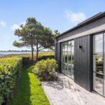 Moderne Villa mit großem Fenster, Blick auf Wasser und Grün. Holzterrasse und Hecke im Vordergrund.