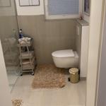 Badezimmer mit Toilette, Fenster und Duschbereich. Fliesenboden und Teppich.