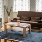 Wohnzimmer mit braunem Sofa, Holztisch und blauem Teppich vor Fenstern mit floralen Vorhängen.