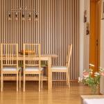 Esszimmer mit Holztisch, vier Stühlen und vertikaler Holzwand. Deckenleuchte mit Glühbirnen.