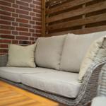 Wicker-Sofa mit Kissen an roter Ziegelwand und Holzgeländer.