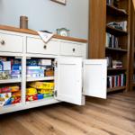 Weißer Schrank mit offenen Türen zeigt Brettspiele. Holzregal und Stuhl im Hintergrund.