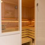 Sauna mit Holzbank und Eimer. Wanduhr und Beleuchtung sichtbar.