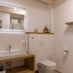Badezimmer mit Waschbecken, Spiegel und Toilette. Holzregal und Dekoration.