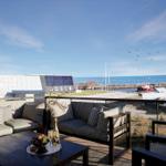 Terrasse mit Sofa, Tisch und Meerblick. Boote liegen am Hafen. Himmel klar, Vögel fliegen.