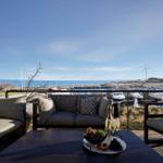 Terrasse mit Sofa und Tisch. Blick auf Meer, Boote und Windkraftanlage.