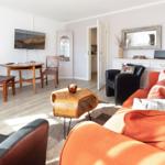 Living room with orange sofa, dining area, and TV. Wooden floor and white walls.