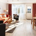 Living area with sofa, armchairs, and dining table. Wooden floor, windows, and door.