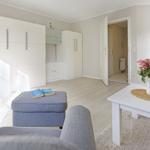 Living area with white cabinets, gray sofa, and kitchen area with flowers.