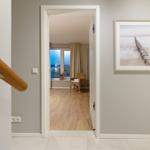 Hausgang mit Holzgeländer und Bild an der Wand. Durch Tür ist Wohnzimmer mit Fenster und Stuhl sichtbar.