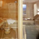 Sauna mit Holzbank, Körbchen und Thermometer. Glaswand zeigt Badebereich.