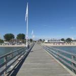 Holzbrücke führt zur Strandpromenade mit Liegestühlen und Gebäuden.
