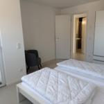Schlafzimmer mit weißem Bett, grauer Sessel und Schrank. Tür führt in Badezimmer.