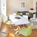 Dining area with white table and green chairs. Living area with sofa and couch. Wooden floor and windows.