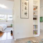 Bedroom with bed and window. Kitchen with white cabinets. Wall with calligraphy and bookshelf.