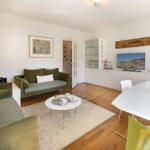 Living and dining area with green sofas, wooden floor, and TV. Table with flowers and side table.