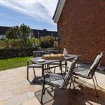 Terrasse mit Tisch und Stühlen neben rotem Backsteinhaus. Grünflächen und Steinmauer im Hintergrund.