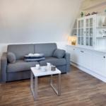 Ein Wohnzimmer mit grauem Sofa, weißem Tisch und weißen Schränken. Holzfußboden und Wandregale.