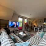 Living area with sofa, TV, and dining area. Windows with garden view.