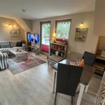 Living and dining area with sofa, TV, table and chairs. Windows with view of greenery.