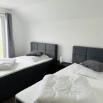 Zwei Betten mit weißen Laken und grauen Kopfkissen. Schwarzer Nachttisch und Fenster mit grauen Vorhängen.