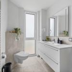 Badezimmer mit Waschtisch, Spiegel, Toilette und Heizung. Fenster sorgt für Tageslicht.