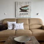 Beige Sofa mit Kissen und Wandbild mit Muschel. Holztisch mit Metallschale.