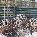 Drei runde Skulpturen aus Hufeisen stehen im Garten vor einem Holzzaun.