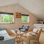 Wohnraum mit Sofa, zwei Sesseln und Schlafcouch unter Dachschrägen. Holzwände und Fenster mit Blick in die Natur.