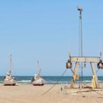 Zwei Fischerboote stehen am Strand. Ein hölzerner Aufbau mit Seilen und einem Mast ist daneben platziert.