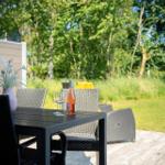 Terrasse mit Tisch, Stühlen und Weinflasche vor grünem Hintergrund.