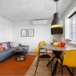 Wohn- und Essbereich mit grauem Sofa, orangefarbenem Teppich und gelben Stühlen um einen runden Tisch.