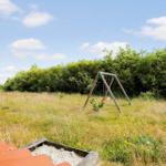Ein Spielplatz mit Schaukel und Sandkasten im Gras. Hintergrund: dichter Busch und blauer Himmel.