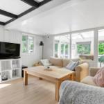 Wohnzimmer mit Sofa, Couchtisch und Fernseher. Große Fenster mit Blick auf Garten.