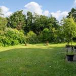 Grüner Garten mit Rasenfläche, Bäumen und einem Haus. Ein kleiner Tisch steht im Zentrum.