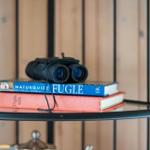 Auf einem Regal liegen Bücher und ein Fernglas. Hintergrund ist Holzwand.