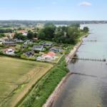 Haus an ruhigem Seeufer mit Bootsanlegestelle und grünen Feldern im Hintergrund.