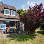 Haus mit Terrasse, Garten und blau-weißem Liegestuhl. Roter Baum und grüne Hecke umgeben den Außenbereich.