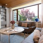 Wohnzimmer mit Sofa, runden Tisch und großen Fenstern. Blick auf Garten mit Bäumen.