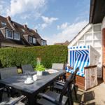 Terrasse mit Tisch, Stühlen und Strandkorb. Hintergrund: grüner Zaun und Häuser.