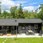 Holzhaus mit Terrasse, Grill und Garten. Umgeben von Bäumen unter blauem Himmel.