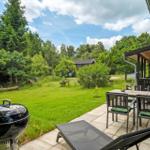 Terrasse mit Grill, Tisch und Stühlen. Grasfläche und Wald im Hintergrund.