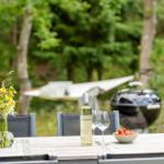 Holztisch mit Wein, Erdbeeren und Blumen im Garten. Grill und Hängematte im Hintergrund.