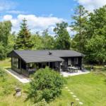 Schwarze Hütte mit Terrasse, Garten und Grillplatz zwischen Bäumen.
