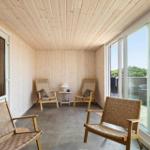 Ein Raum mit Holzwänden und -decke, zwei Sessel und einem Tisch. Große Fenster bieten Blick auf Außenbereich.