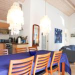 Essbereich mit blauer Tischdecke und Holzstühlen unter hängenden Lampen. Wohnbereich mit Sofa und Wandbild.