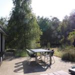 Terrasse mit Tisch, Stühlen und Sonnenschirm. Hintergrund: Wald. Links: Gebäude mit Glasfront.