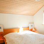 Schlafzimmer mit Holzdecke, Bett und Fenster. Auf Nachttischen stehen Lampen und Bilder.