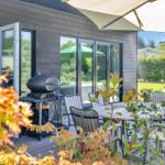 Terrace with table, chairs, umbrella, and grill. Background: garden with green areas and trees.