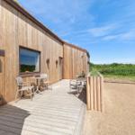 Holzhaus mit Terrasse, Tisch und Stühlen. Grünflächen im Hintergrund.